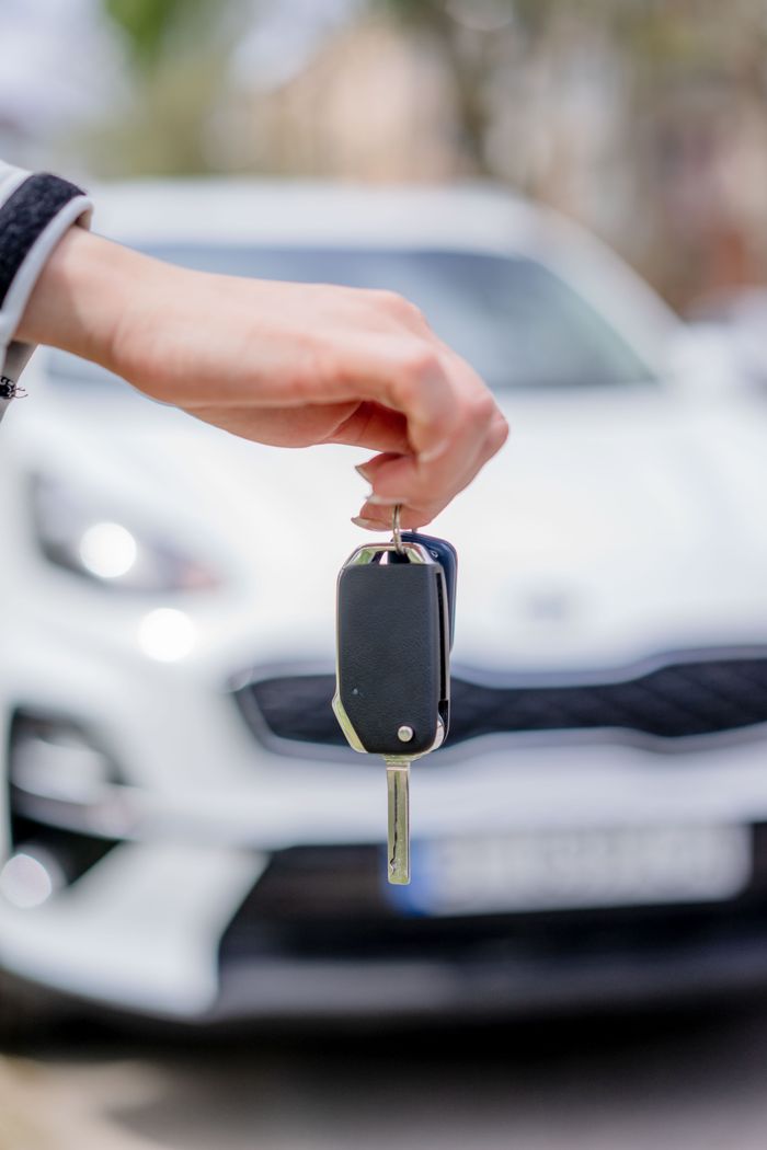 Services Close-up of a hand holding a car key, with a white car in the background outdoors.